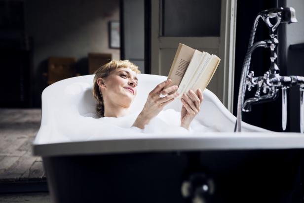 Eine Frau entspannt in einer mit Schaum gefüllten Badewanne und liest ein Buch.