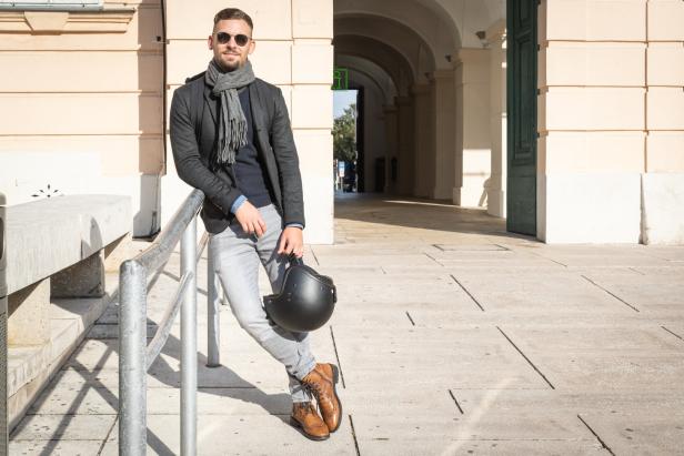 Ein Mann mit Sonnenbrille lehnt an einem Geländer und hält einen Motorradhelm.