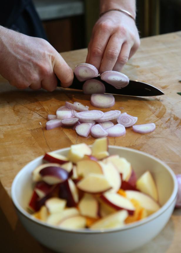 Eine Person schneidet Schalotten auf einem Holzbrett, im Vordergrund eine Schüssel mit Obststücken.