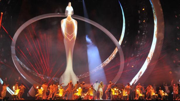 Eine Sängerin steht mit Tänzern und Schlagzeugern auf einer Bühne vor einer großen Statue.