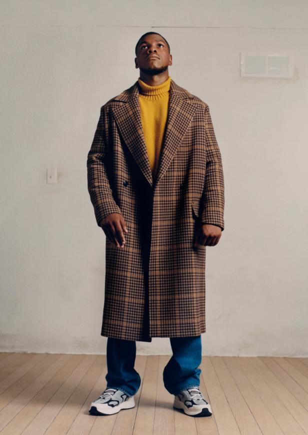 Ein Mann mit Mantel, Rollkragenpullover und Jeans steht auf einem Holzboden.