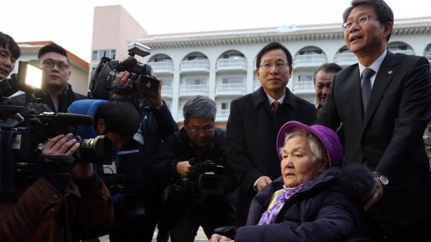 Eine ältere Frau im Rollstuhl wird von einer Gruppe von Reportern und Beamten umgeben.