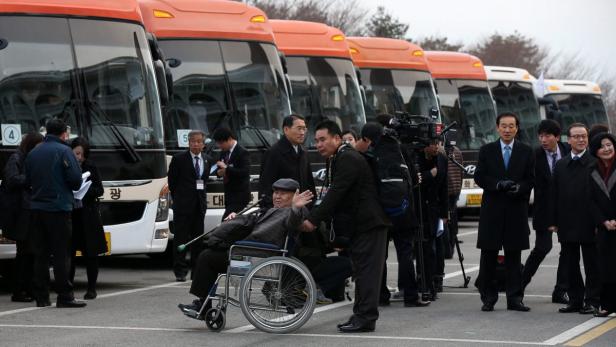 Eine Gruppe von Menschen steht vor einer Reihe orangefarbener Busse, eine Person sitzt im Rollstuhl.
