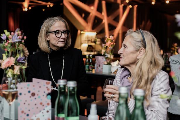 Zwei Frauen unterhalten sich bei einer Veranstaltung mit Blumen und Getränken.