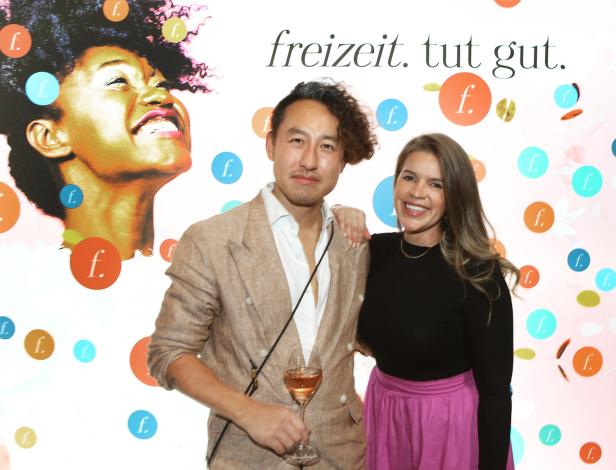 Ein Mann im Anzug und eine Frau posieren vor einer Wand mit dem Schriftzug „freizeit. tut gut.“.