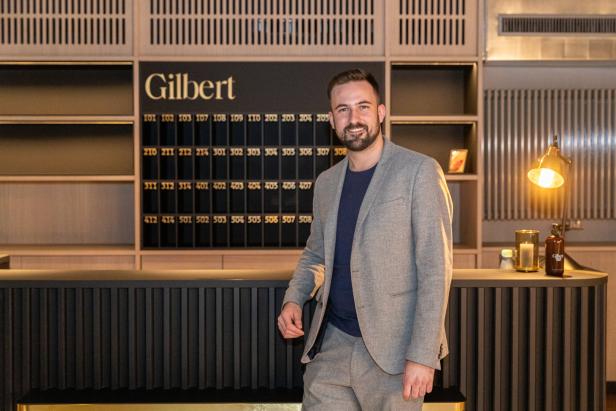 Ein Mann in einem grauen Anzug steht vor der Rezeption des Hotels „Gilbert“.