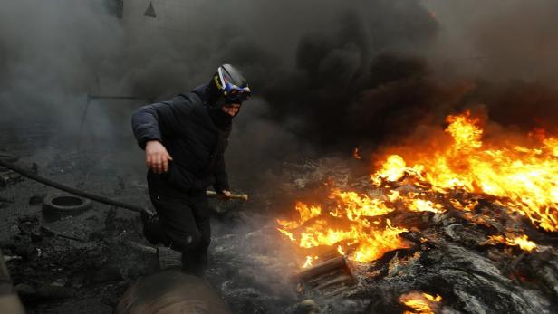 Eine Person mit Schutzbrille rennt an einem großen Feuer vorbei, umgeben von Rauch.