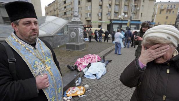 Ein orthodoxer Priester steht vor zwei Leichen, während eine Frau ihr Gesicht verdeckt.
