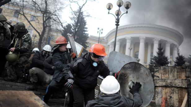 Demonstranten in Schutzkleidung und Helmen stehen vor einem Gebäude mit Säulen.