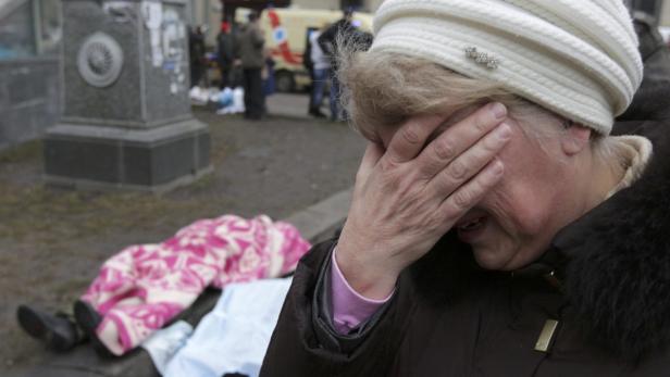 Eine weinende Frau steht neben einer auf dem Boden liegenden Person, die mit einer Decke bedeckt ist.