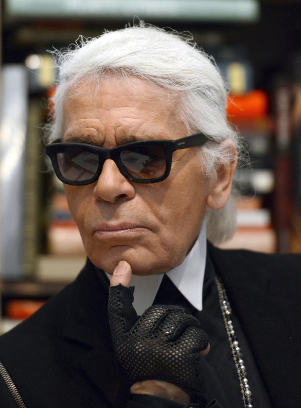 Karl Lagerfeld mit Sonnenbrille und schwarzem Anzug posiert mit einer Hand am Kinn.