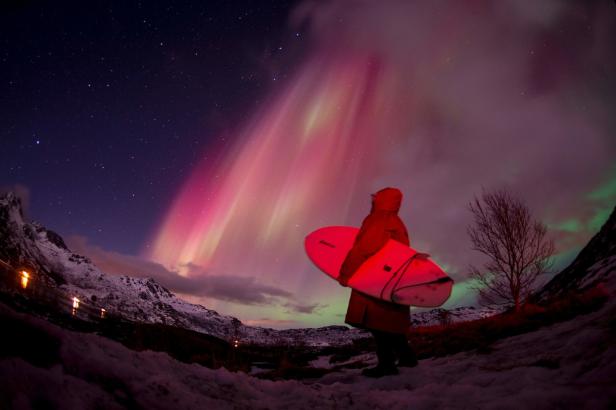 Eine Person mit Surfbrett beobachtet die Nordlichter über einer verschneiten Landschaft.