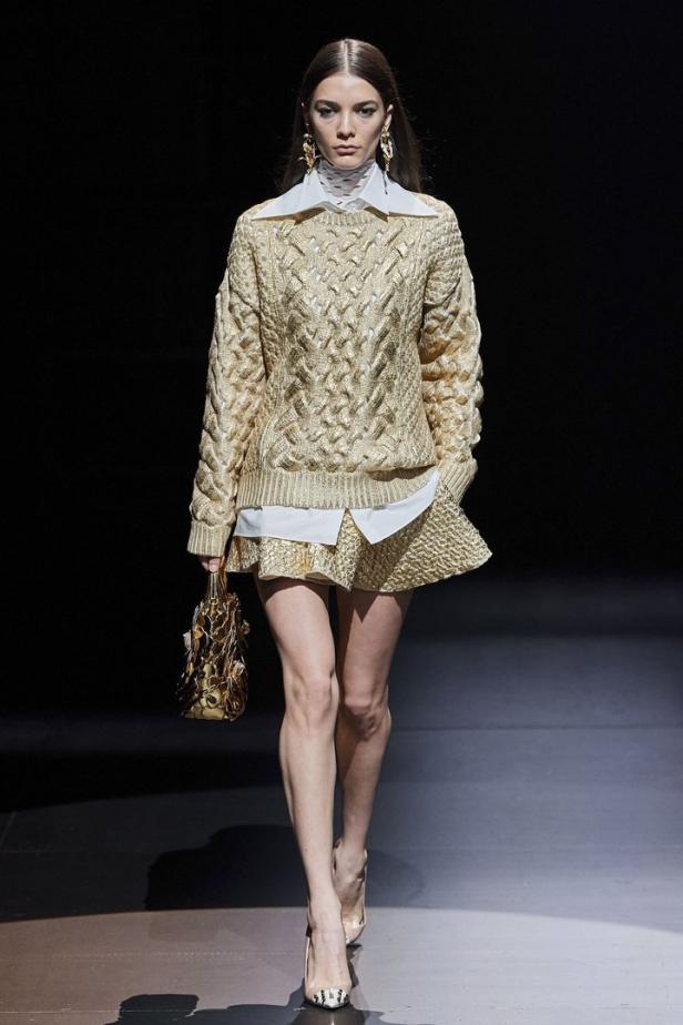 Eine Frau in einem goldenen Strickpullover und Rock auf einem Laufsteg.