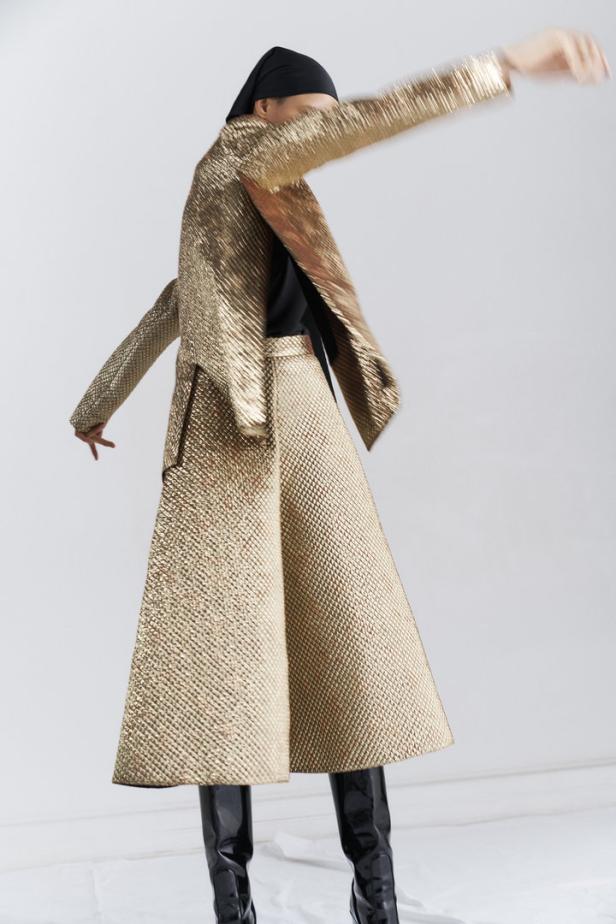 Eine Person in einem goldenen Blazer und Rock mit schwarzen Stiefeln vor einem weißen Hintergrund.