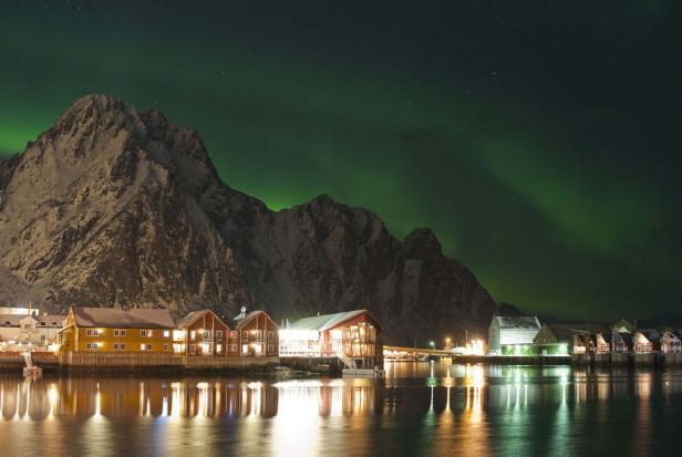 Nordlichter über einem Küstenort mit schneebedeckten Bergen im Hintergrund.