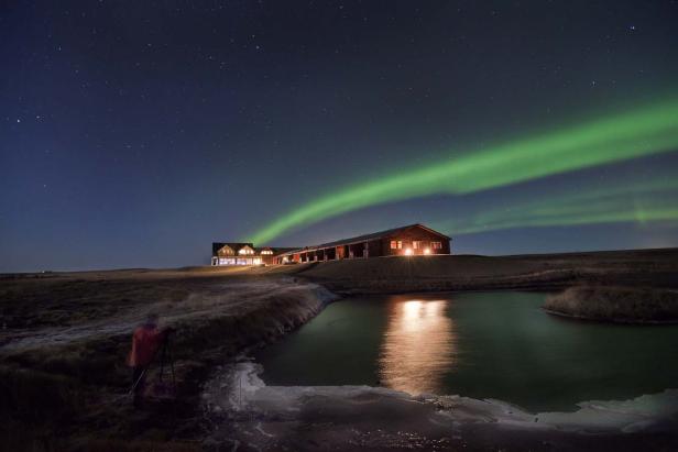 Ein Holzhaus unter dem Nachthimmel mit grünem Polarlicht und einem kleinen See im Vordergrund.