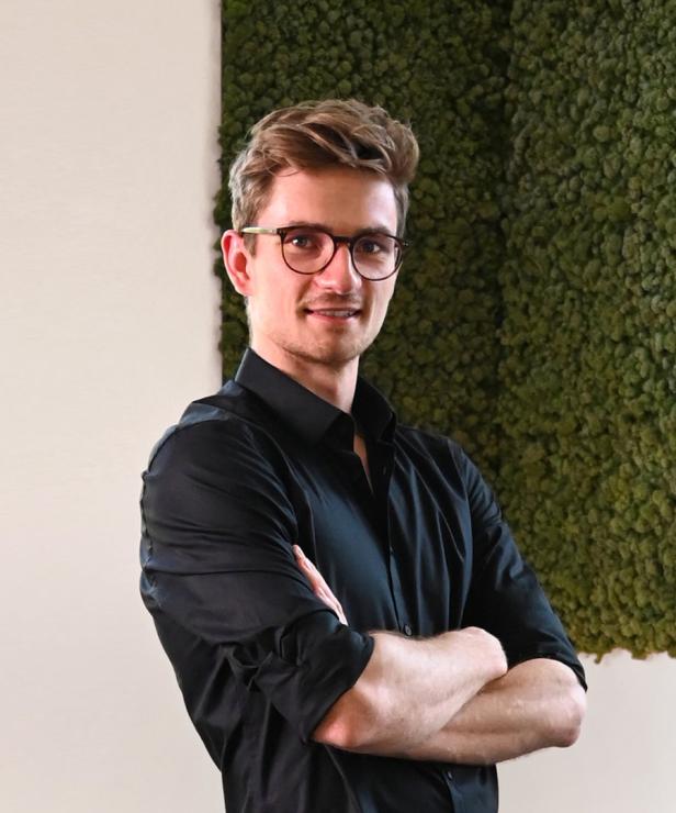 Ein junger Mann mit Brille und schwarzem Hemd steht mit verschränkten Armen vor einer grünen Wand.