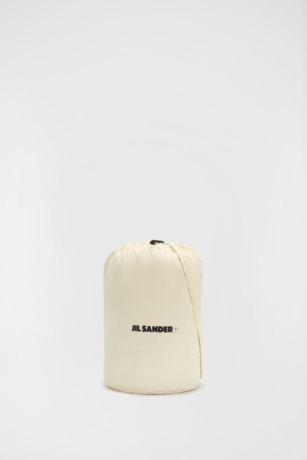 Ein beiger Packsack mit dem Logo von Jil Sander+ steht auf einem weißen Hintergrund.