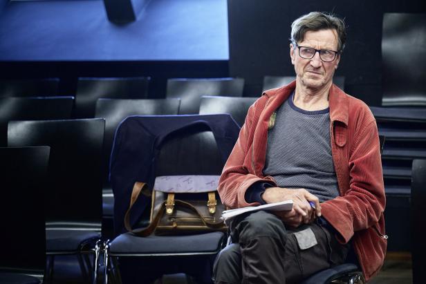 Ein Mann mit Brille und roter Jacke sitzt in einem leeren Zuschauerraum.
