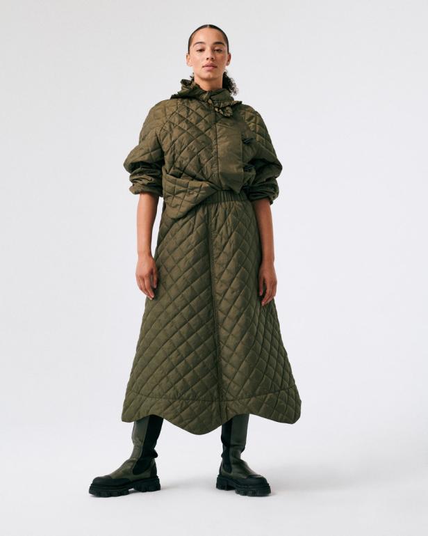 Eine Frau trägt ein gestepptes, olivgrünes Outfit, bestehend aus Jacke und Rock, mit schwarzen Stiefeln.