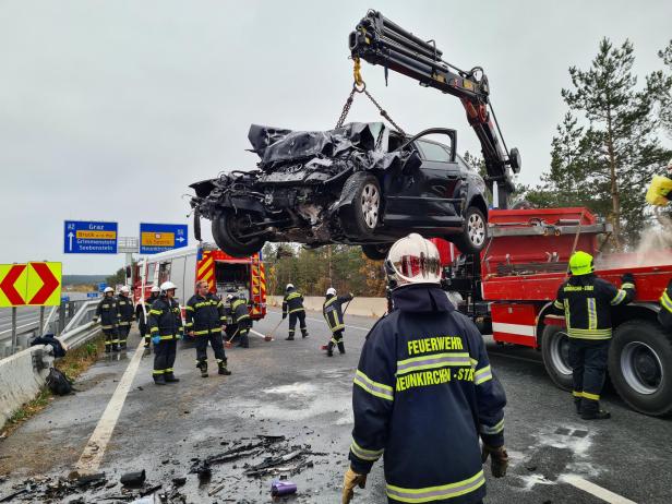 Ein schwer beschädigtes Auto wird von einem Kran auf einer Autobahn angehoben, während Feuerwehrleute die Straße reinigen.