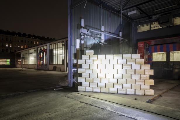 Eine Mauer aus hellen Ziegelsteinen steht vor einer Industriehalle.
