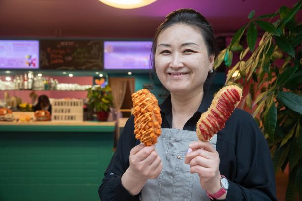 Eine lächelnde Frau präsentiert zwei koreanische Corn Dogs in einem Restaurant.