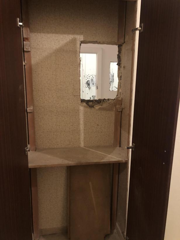Ein offener Kleiderschrank mit einem Loch in der Rückwand, das einen Blick in einen anderen Raum freigibt.