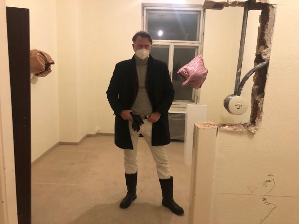 Ein Mann mit Maske, Mantel und Stiefeln posiert in einem unrenovierten Raum mit Kunstobjekten an den Wänden.