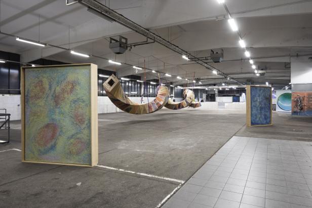 In einer Lagerhalle hängen zwei skulpturale Objekte aus Karton und zwei gerahmte Gemälde stehen auf dem Boden.
