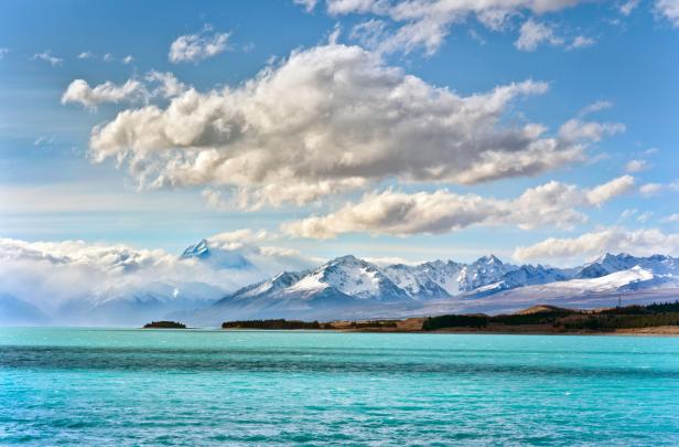 Türkisfarbenes Wasser unter einem Himmel mit Wolken und schneebedeckten Bergen.