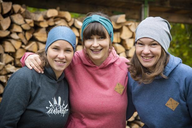 Drei lächelnde Frauen mit Stirnbändern und Mützen stehen vor einem Holzstapel.