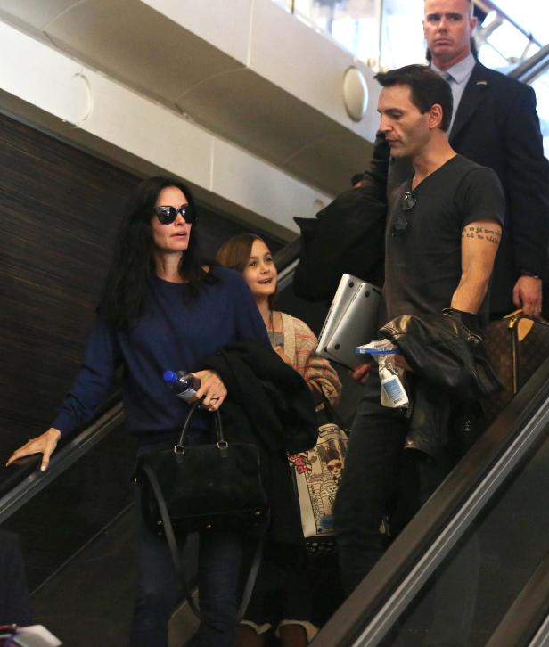 Courteney Cox mit Familie auf einer Rolltreppe in einem Flughafen.