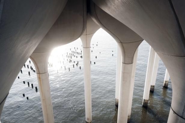 Blick von unten auf die Pfeiler einer Brücke, die sich im Wasser spiegeln.