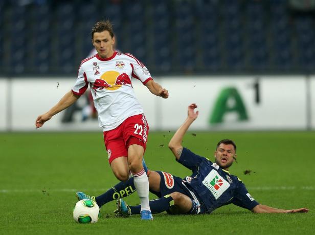 Ein Fußballspieler in Rot-Weiß dribbelt den Ball, während ein Gegenspieler zu Boden geht.