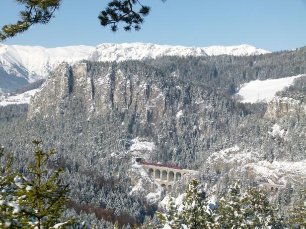 Ein roter Zug fährt über ein schneebedecktes Viadukt in einer winterlichen Berglandschaft.