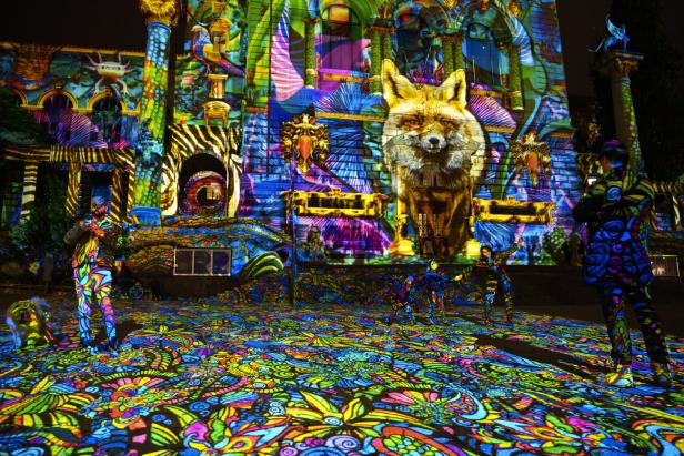 Eine farbenfrohe Lichtprojektion zeigt ein Gebäude mit einem Fuchs und Menschen in gemusterten Anzügen.