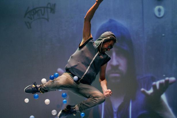 Ein Mann mit Kapuzenweste springt vor einer Projektion mit blauen und weißen Bällen.