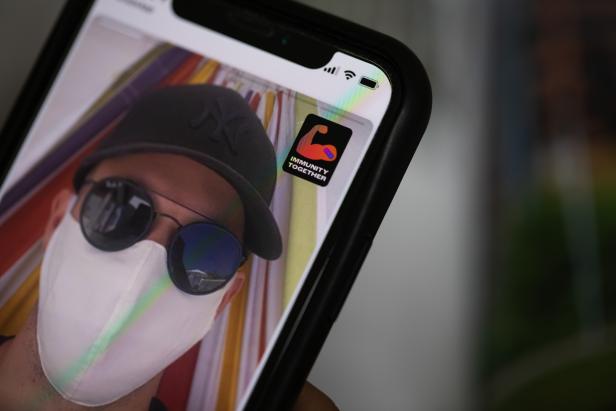 Auf dem Bildschirm eines Smartphones ist ein Mann mit Maske und Kappe zu sehen.