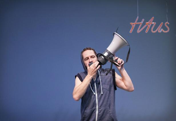 Ein Mann mit Kapuzenweste spricht in ein Megafon vor einem blauen Hintergrund mit der Aufschrift „Titus“.