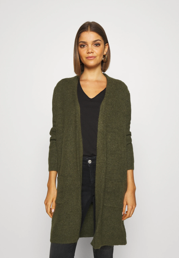 Eine Frau trägt eine lange, olivgrüne Strickjacke über einem schwarzen T-Shirt und Jeans.