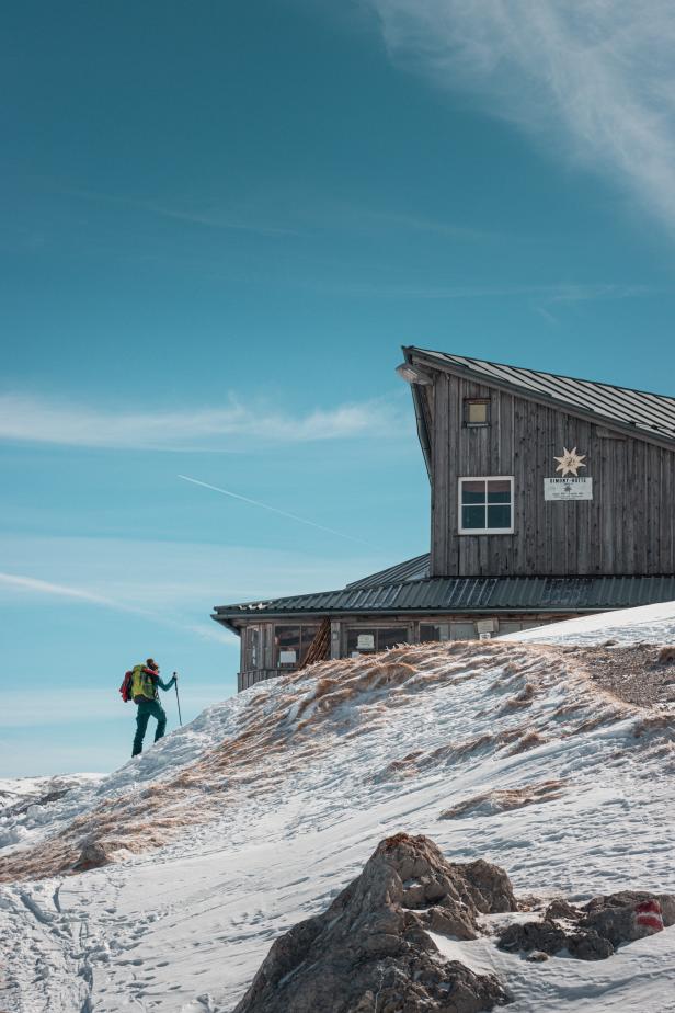 Eine Wanderin steigt mit Rucksack und Stöcken auf eine schneebedeckte Hütte zu.