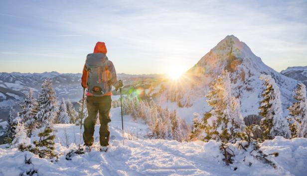 Eine Person mit Rucksack blickt auf eine verschneite Berglandschaft im Sonnenlicht.