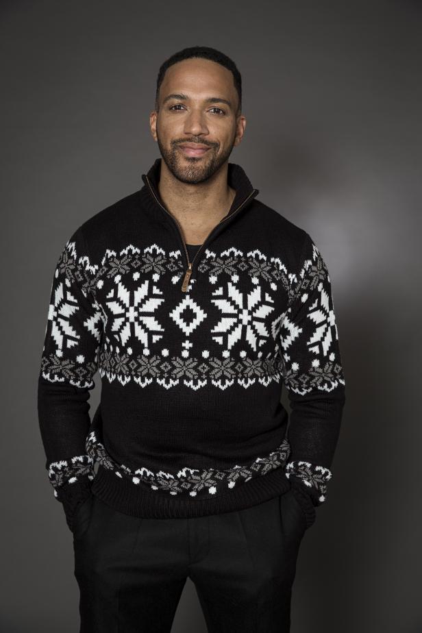 Ein Mann mit Bart und einem Norwegerpullover steht vor einem grauen Hintergrund.