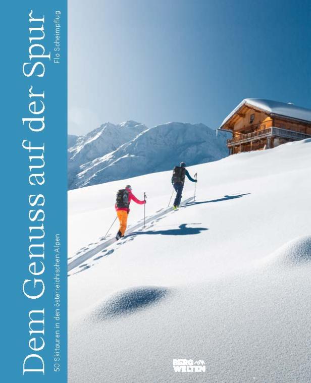 Zwei Skitourengeher steigen im Schnee bergauf, im Hintergrund eine Hütte und Berge.