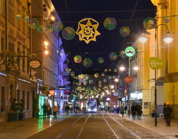 Eine abendliche Szene in Linz mit Weihnachtsbeleuchtung über einer belebten Straße mit Straßenbahnschienen.