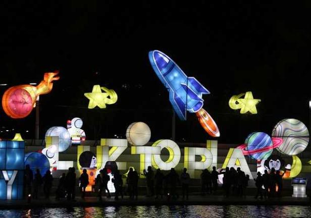 Illuminierte Raumfahrt-Dekorationen und der Schriftzug „Luztopia“ spiegeln sich im Wasser.
