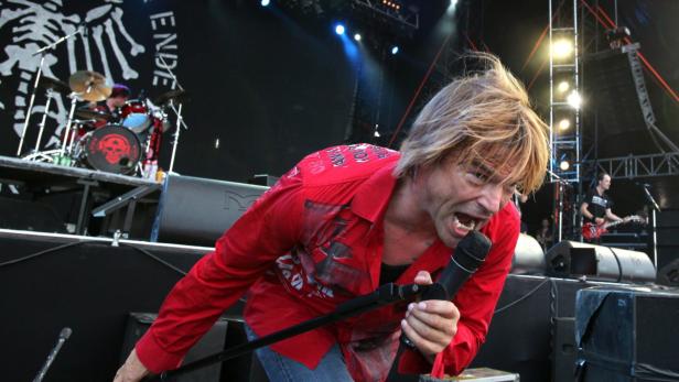 Ein Sänger mit rotem Hemd schreit in ein Mikrofon auf einer Bühne.