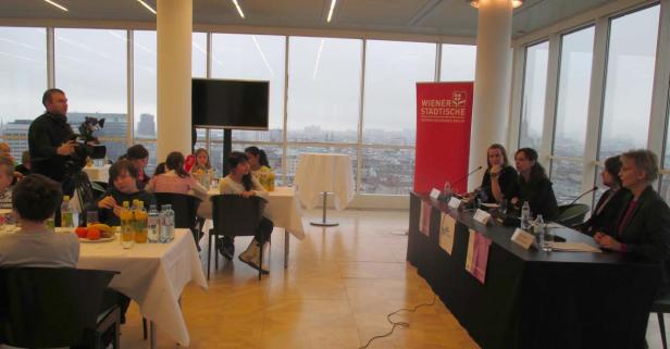 In einem Raum mit Blick auf Wien sitzen Kinder an Tischen, während eine Podiumsdiskussion stattfindet.