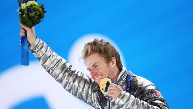 Ein strahlender US-amerikanischer Athlet mit Goldmedaille und Blumenstrauß vor blauem Hintergrund.